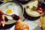 Hand mit Löffel Marmelade neben Frühstücksgerichten.