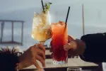 Zwei Hände stoßen farbige Cocktails in einer malerischen Umgebung an