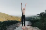 Person mit erhobenen Armen auf einem Felsen mit Bergen im Hintergrund