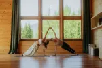 Zwei Personen machen Yoga in einem Raum mit großen Fenstern.