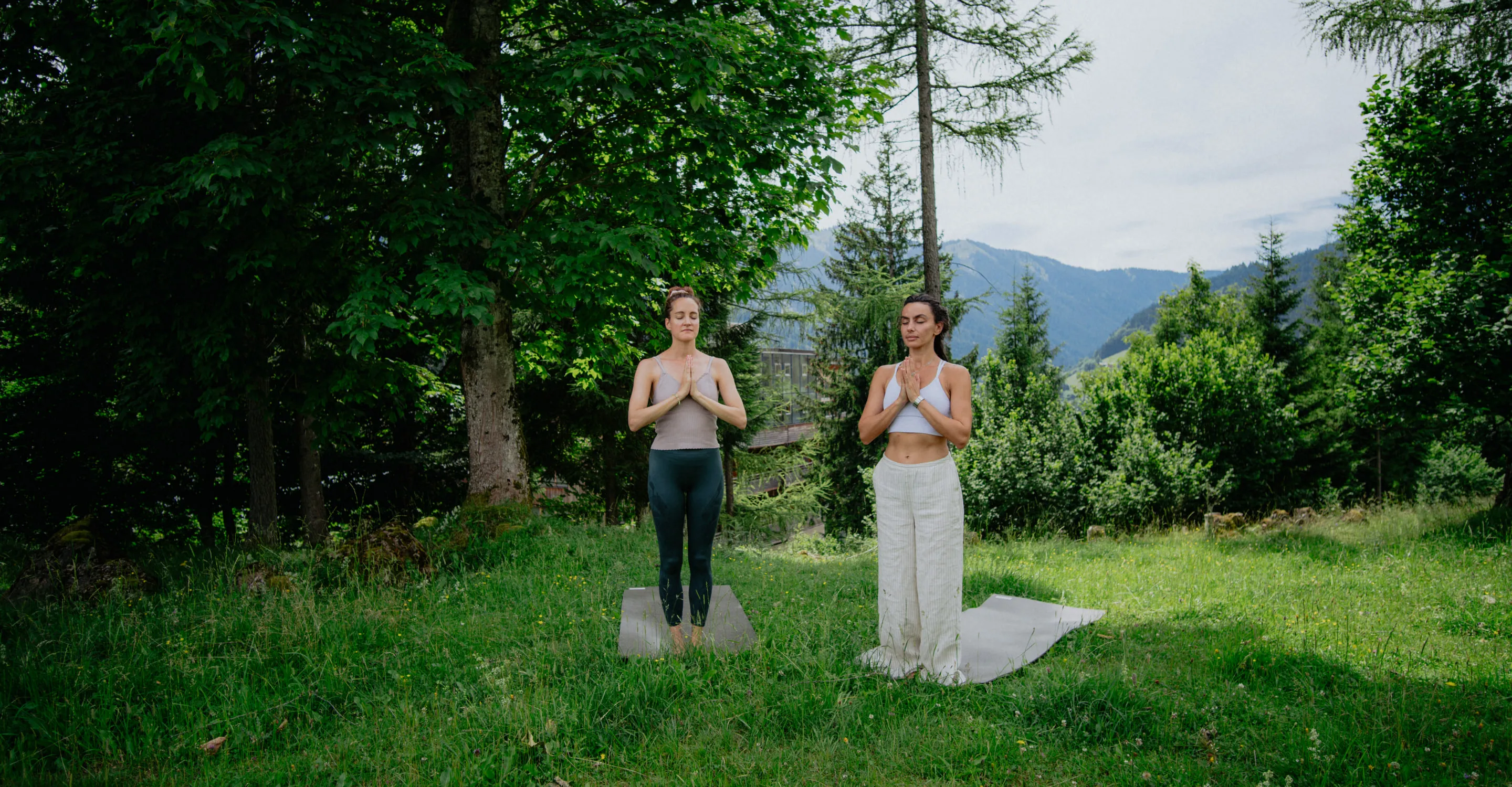 Zwei Frauen praktizieren Yoga im Freien zwischen Bäumen und Bergen.
