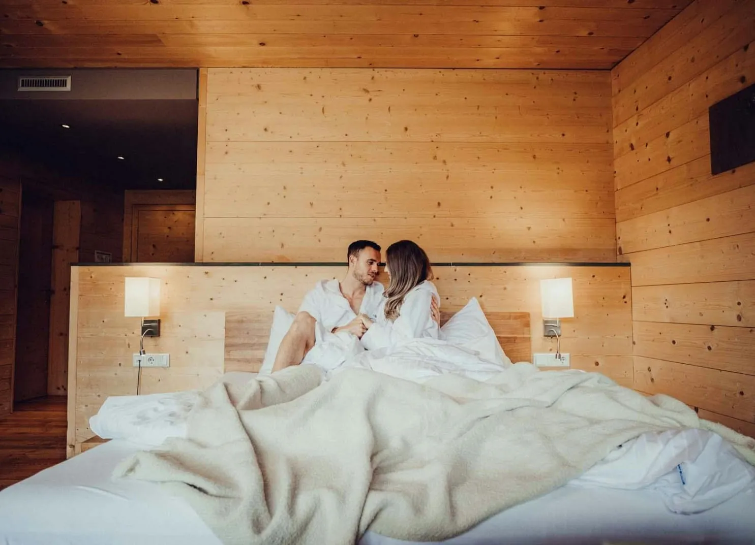 Paar sitzt auf einem Bett in einem gemütlichen Holzraum und lächelt sich an
