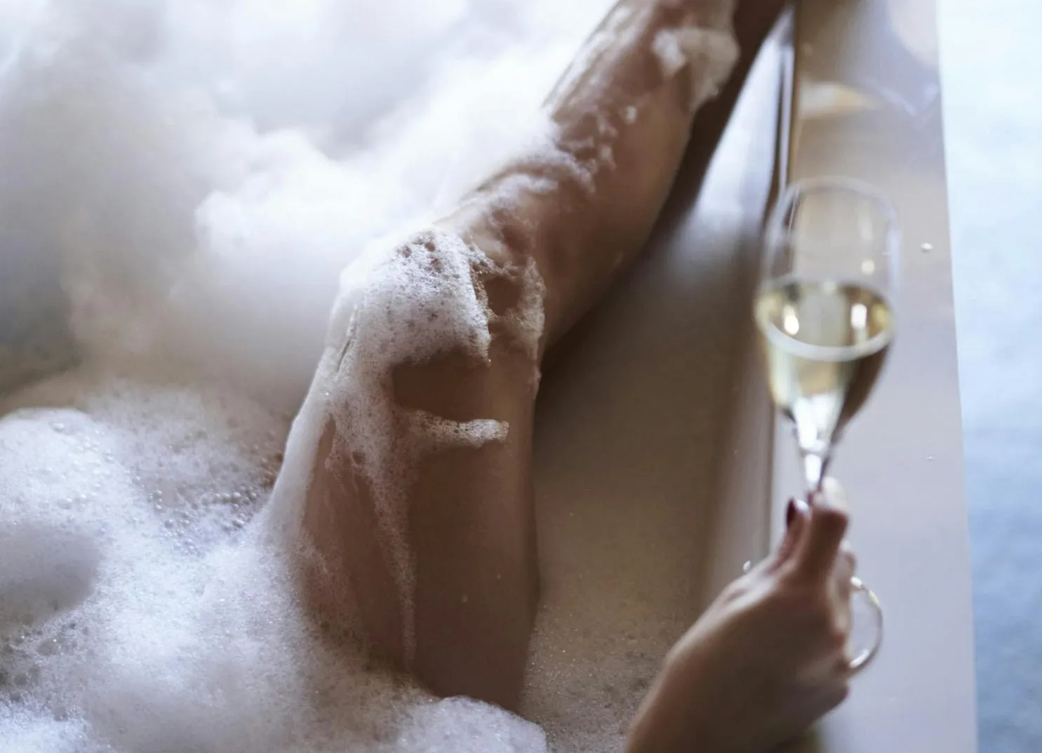 Eine Hand, die ein Glas Sekt neben einem Bein in einem Schaumbad hält