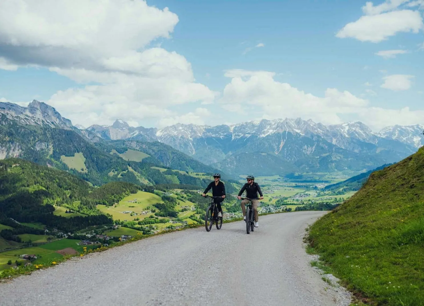 Zwei Radfahrer auf einer Straße, umgeben von Bergen und grüner Landschaft.