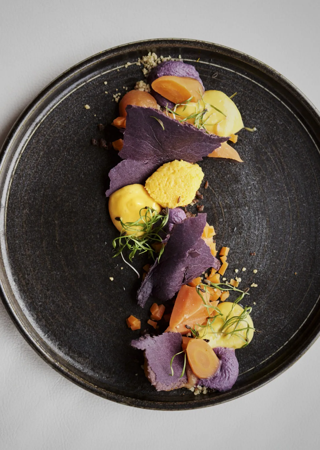 Gericht mit lila Chips, gelber und oranger Creme, dekoriert mit grünen Kräutern.