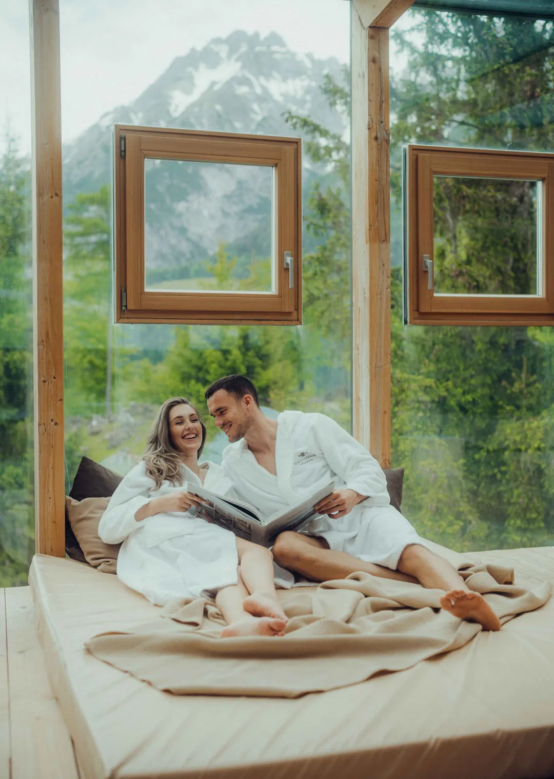 Pärchen in Bademänteln sitzen auf einem Bett mit Blick auf Berge