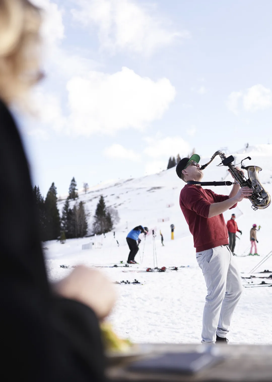 Mann mit Saxophon auf einem schneebedeckten Hügel, im Hintergrund Skifahrer.