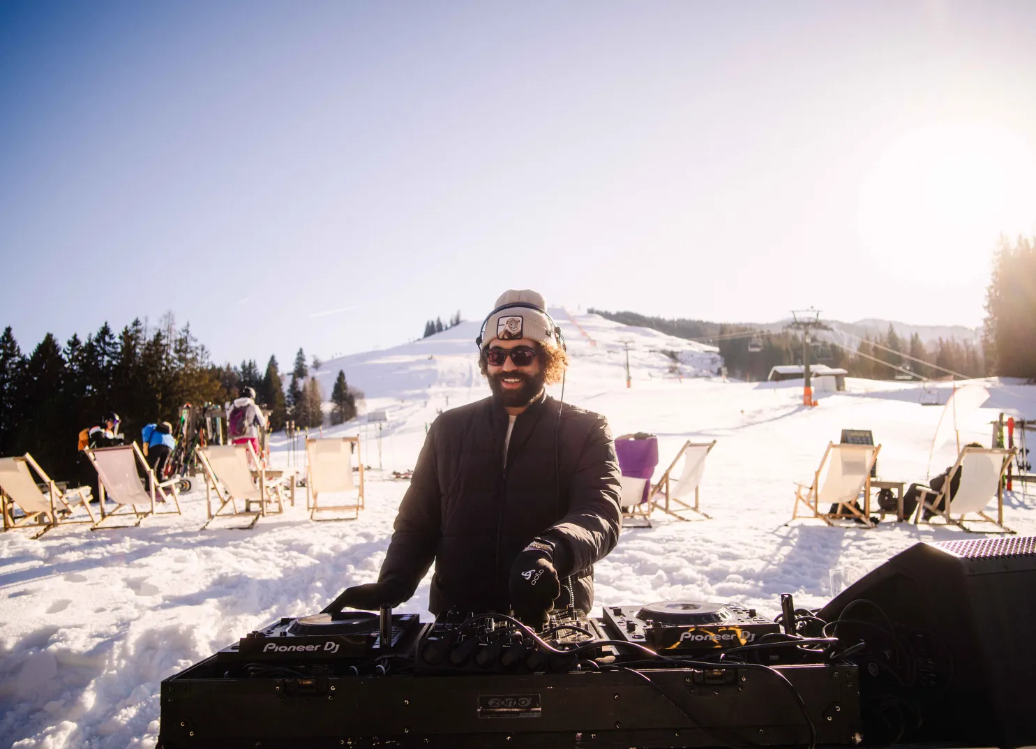 DJ steht an einem Mischpult auf einer verschneiten Terrasse mit Gästen im Hintergrund.